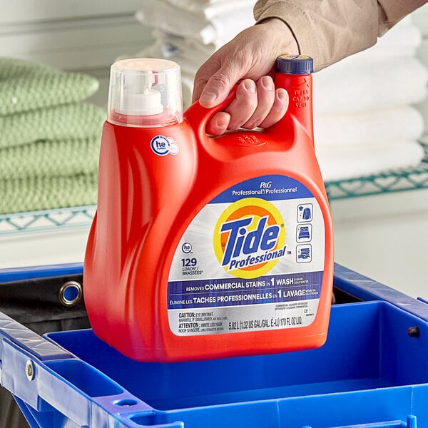A person's hand holding a red bottle of Tide Professional laundry detergent in a blue basket.