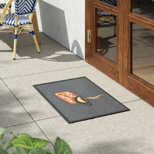 A rectangular entrance mat with a 'Hot Coffee' design placed outside a wooden door on a tiled patio.