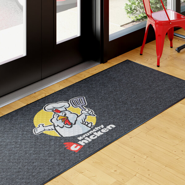 A rectangular black entrance mat with a cartoon chicken chef and the text 'krunchy chicken,' placed near a glass door on a wooden floor.