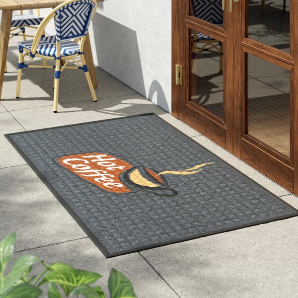 A rectangular gray entrance mat with a waffle texture and a 'Hot Coffee' design, placed outside a door.