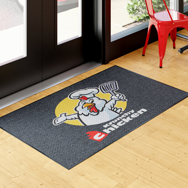 A rectangular entrance mat with a cartoon chicken chef and the text 'Krunchy Chicken' printed on it, placed indoors near a door.