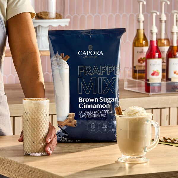 A bag of Capora Brown Sugar Cinnamon Frappe Mix displayed on a counter with two prepared drinks and syrup bottles in the background.