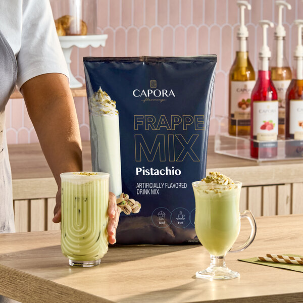 A bag of Capora Pistachio Frappe Mix is displayed on a counter with two glasses of prepared pistachio frappe drinks.
