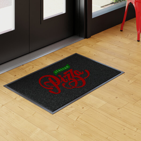 A rectangular black entrance mat with red and green lettering that reads 'Italian Pizza,' placed on a light wood floor near a glass door.