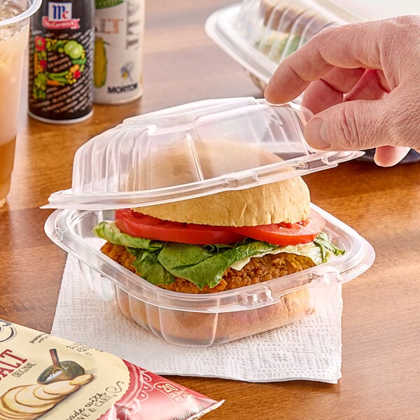 A hand holding a burger in a Dart clear hinged plastic container.