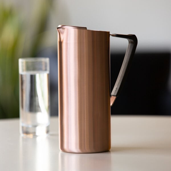 A slim rose gold stainless steel pitcher with a black handle and a guard, placed on a table next to a glass of water.