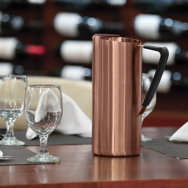 A slim rose gold stainless steel pitcher with a black handle and guard, placed on a set dining table.