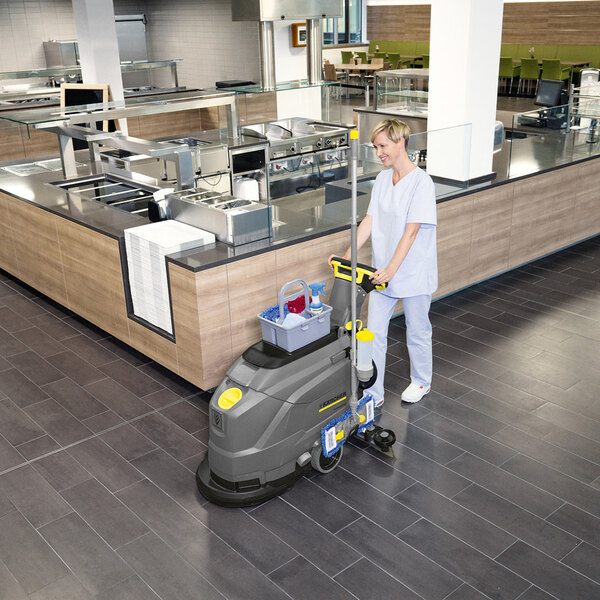 A person operating a Karcher BD 43/25 C BP 17-inch cordless walk-behind floor scrubber in a commercial kitchen or cafeteria setting.
