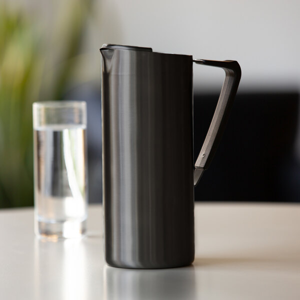 A slim black onyx stainless steel pitcher with a handle and spout, placed on a table next to a glass of water.