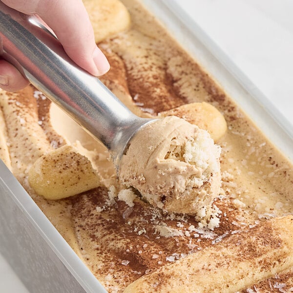 A hand scooping Villa Dolce Tiramisu Gelato into a metal pan on a counter.