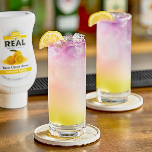 Two glasses of pink and yellow drinks on a table with a bottle of Real Yuzu Puree Infused Syrup.