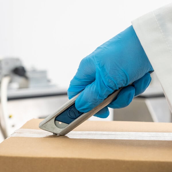 A person in a blue glove using a Pacific Handy Cutter metal-detectable safety knife to open a box.