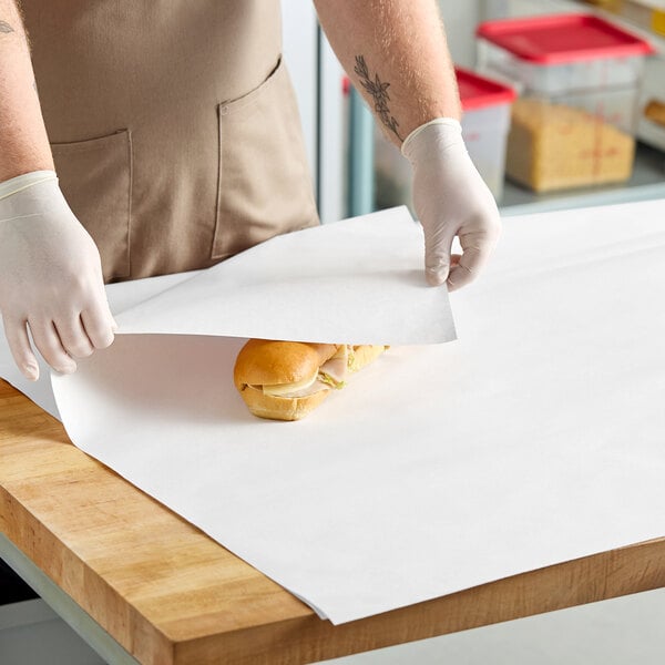 A large sheet of white newsprint sandwich wrap paper being used to wrap a sandwich on a wooden countertop.