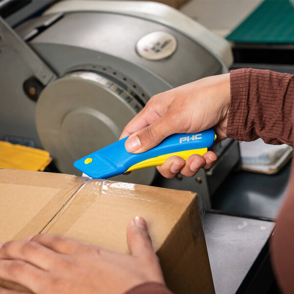 A person using a Pacific Handy Cutter blue and yellow safety knife to cut a box.