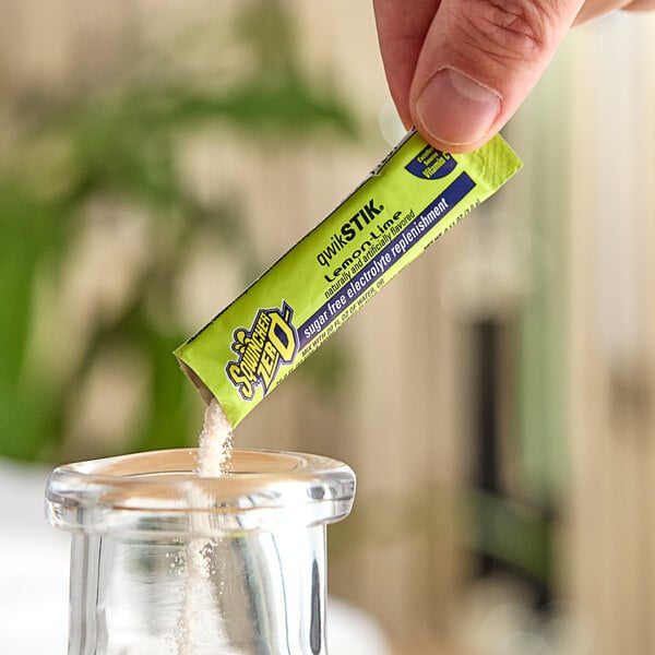 A hand holding a Sqwincher QwikStik Zero Lemon-Lime packet over a glass container.