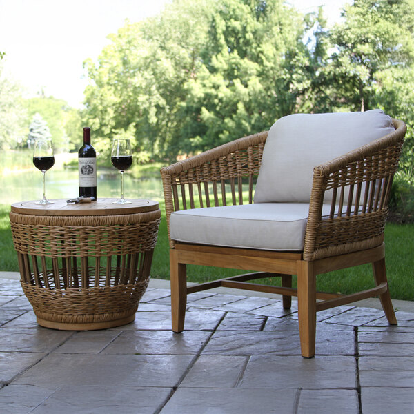 A wicker table with a wicker basket and wine glasses on top next to a bottle of wine on a stone patio.