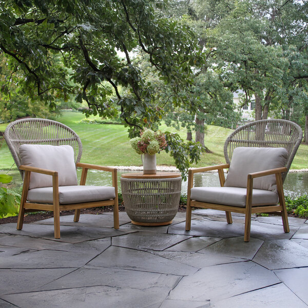 An Outdoor Interiors teak and wicker storage table on a stone patio with a vase of flowers on it.