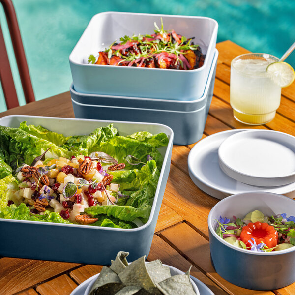 A group of Cal-Mil Stone Blue square Bento Boxes on a table with bowls of food.