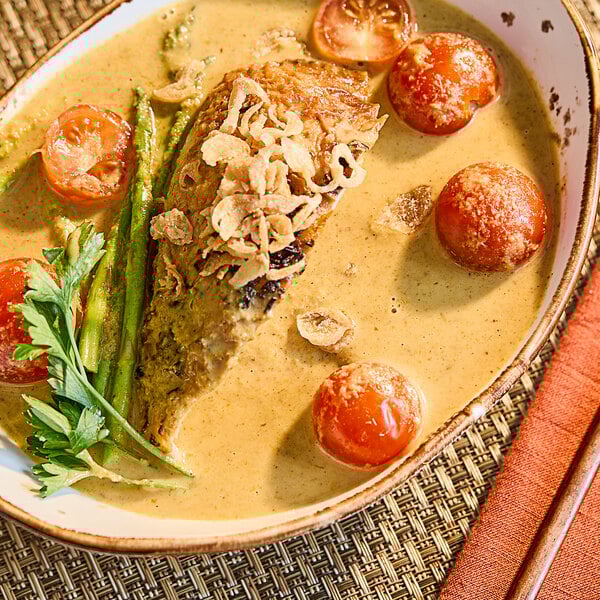 A bowl of yellow curry with a piece of fish, cherry tomatoes, asparagus, fried shallots, and fresh herbs.