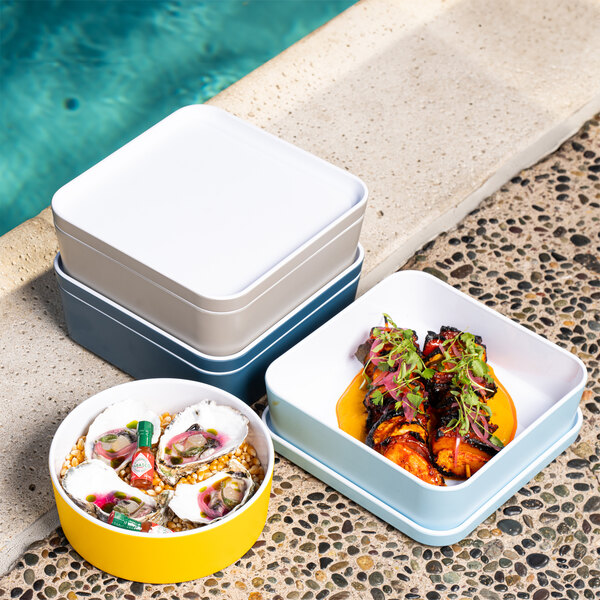 Three Cal-Mil ocean blue square melamine bento boxes filled with food on a table near a pool.
