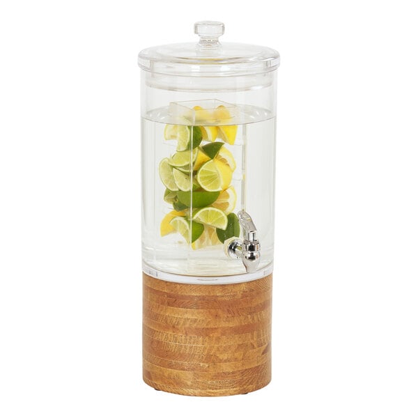 A Cal-Mil round glass beverage dispenser with lemons and limes in it on a rustic pine base.