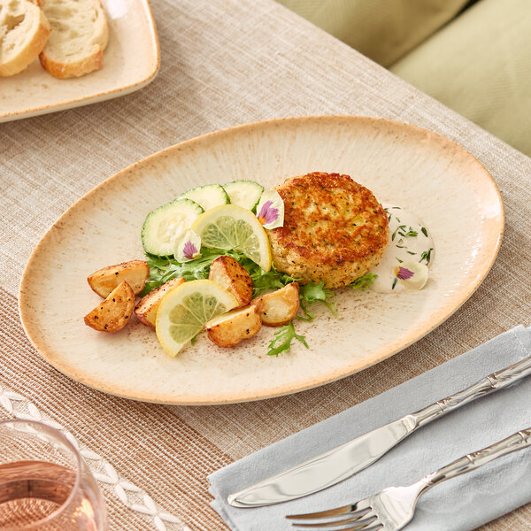 An oval beige porcelain platter displaying a meal with potatoes, lemon slices, cucumber, and a crab cake.