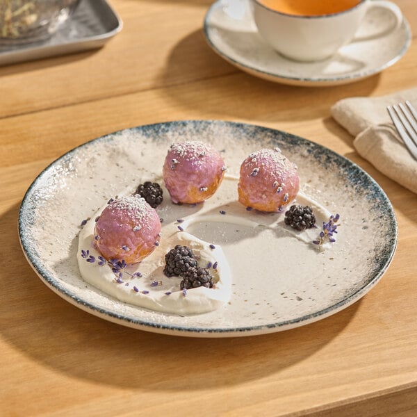 A round gray porcelain coupe plate with a speckled rim, featuring a dessert with cream, blackberries, and purple-glazed pastries.