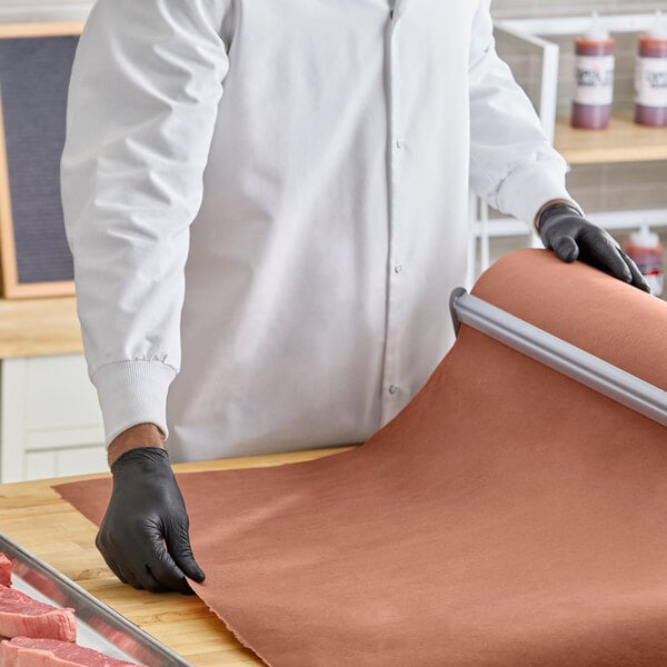 A roll of pink or peach butcher paper being unrolled on a countertop by a person wearing black gloves and a white coat.