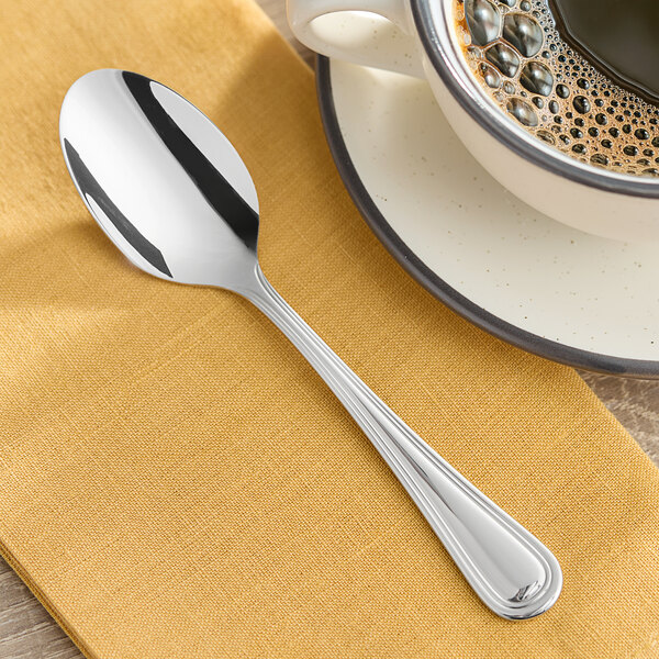 An Acopa Edgeworth stainless steel teaspoon on a saucer next to a cup of coffee.