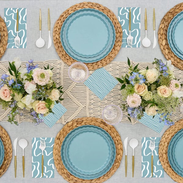 A table set with blue Sophistiplate Simply Eco pool plant fiber dinner plates and flowers on a woven mat.