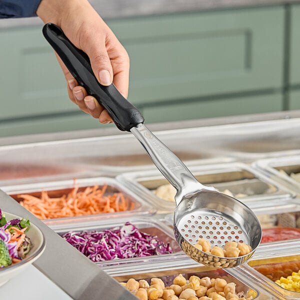 A hand using a Vollrath black perforated round Spoodle over a tray of food.
