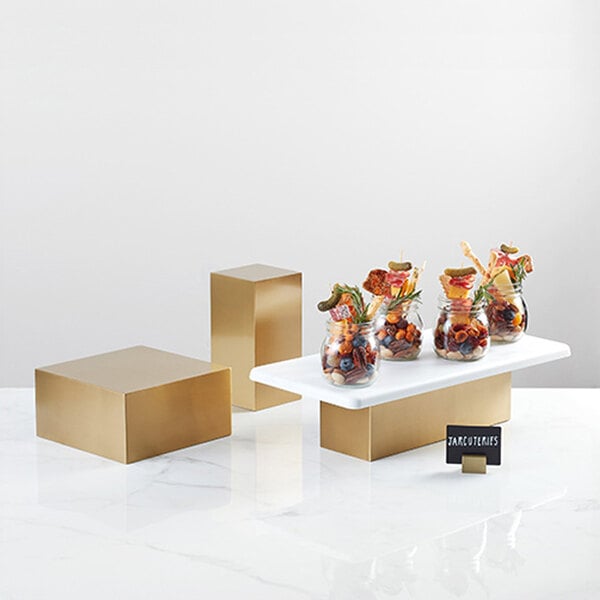 An American Metalcraft gold stainless steel rectangular riser on a white surface with a jar of dried fruits and nuts inside.