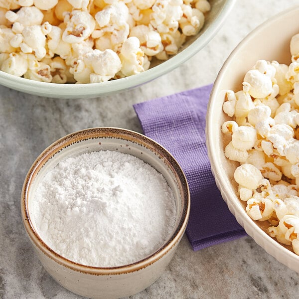 A bowl of white powdered sweet and salty popcorn seasoning next to bowls of popcorn.