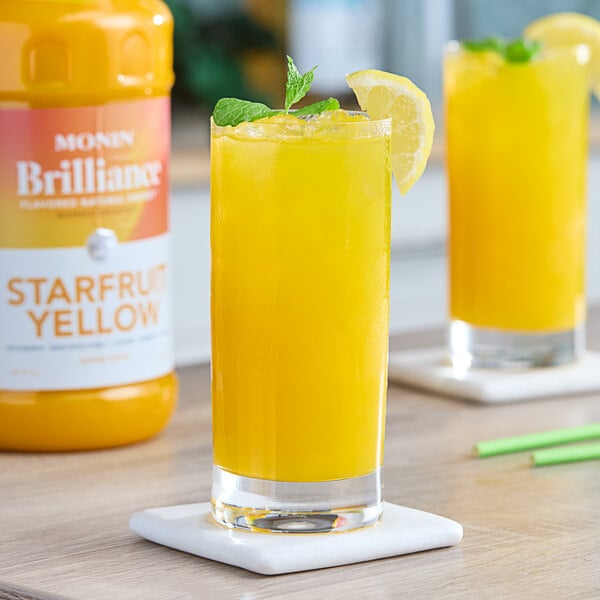 A close up of a glass of yellow Monin Brilliance Starfruit Natural Energy drink with lemon slices and mint leaves.