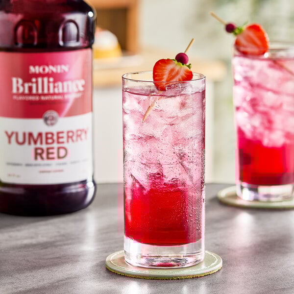 A tall glass of iced red beverage garnished with a strawberry and berry skewer, with a bottle of Monin Brilliance Yumberry Red Natural Energy Concentrate in the background.