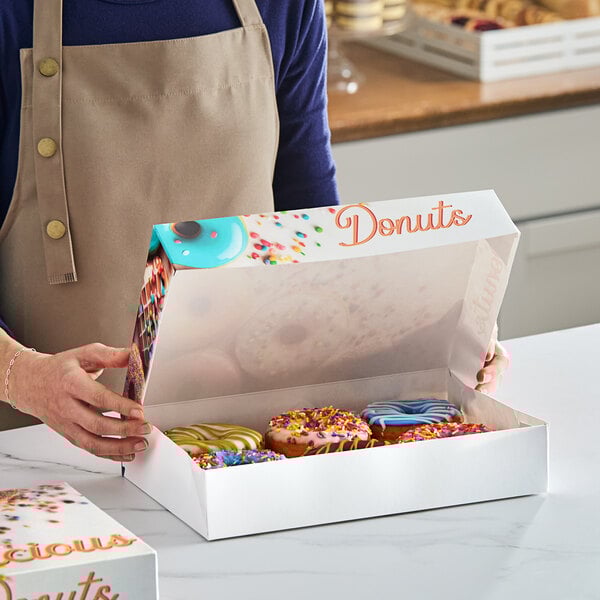 A woman holding a white "Delicious" donut box on a bakery counter.