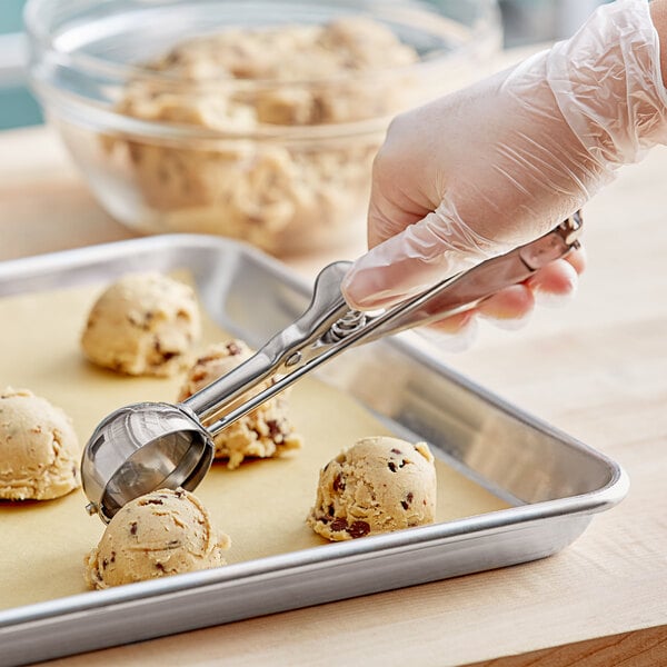 A stainless steel squeeze handle disher being used to scoop cookie dough onto a baking tray.