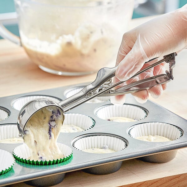 A stainless steel squeeze handle disher being used to portion batter into cupcake liners in a muffin tin.