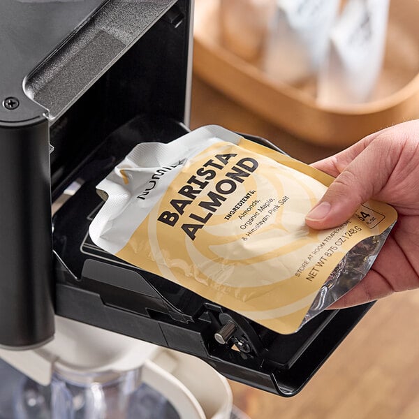 A person holding a package of Numilk Barista Almond Milk in front of a coffee machine.