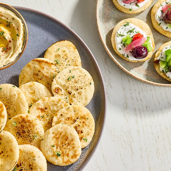A plate of small, round pre-grilled cocktail flatbreads served with a dip and assorted toppings.