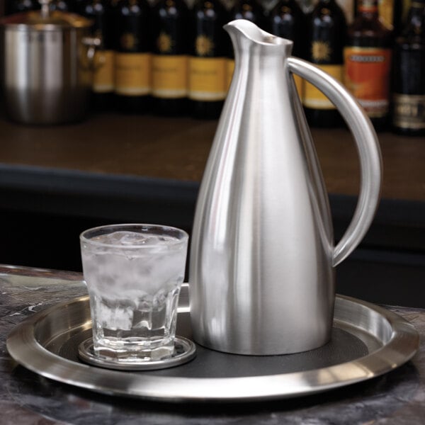 A silver jug and a glass of ice on a round stainless steel non-skid serving tray.