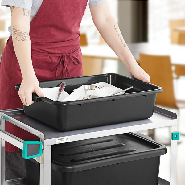 A black polypropylene bus tub filled with used dishes and utensils on a cart in a restaurant setting.