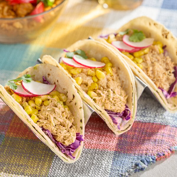 A trio of tacos filled with plant-based vegan tuna, corn, shredded cabbage, and sliced radishes on a colorful cloth.