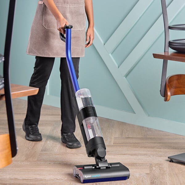 A person using a Dyson WashG1 cordless wet and dry vacuum cleaner on a wooden floor.