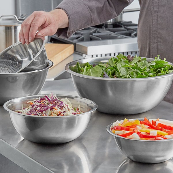 A set of stainless steel mixing bowls in various sizes, some filled with salad and chopped vegetables, on a kitchen counter.