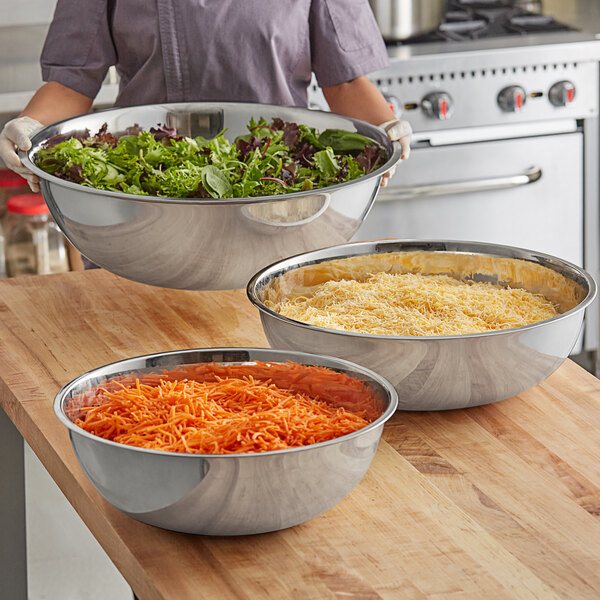 A set of three extra-large stainless steel mixing bowls filled with various ingredients on a wooden countertop.