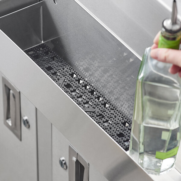 A hand using a Choice black plastic interlocking bar mat strip to hold a green bottle over a stainless steel sink.