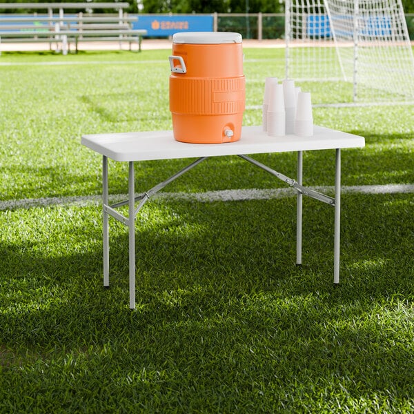 A rectangular white plastic folding table with gray metal legs set up on a grassy field, holding an orange beverage cooler and a stack of white cups.