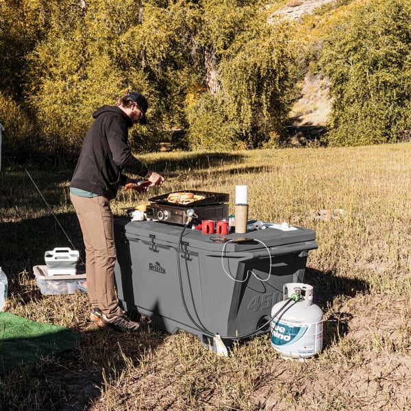A man cooking food on a grill next to a large grey Grizzly outdoor cooler.