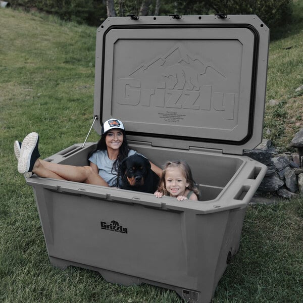 A woman and two children sitting in a Grizzly outdoor cooler with a dog.
